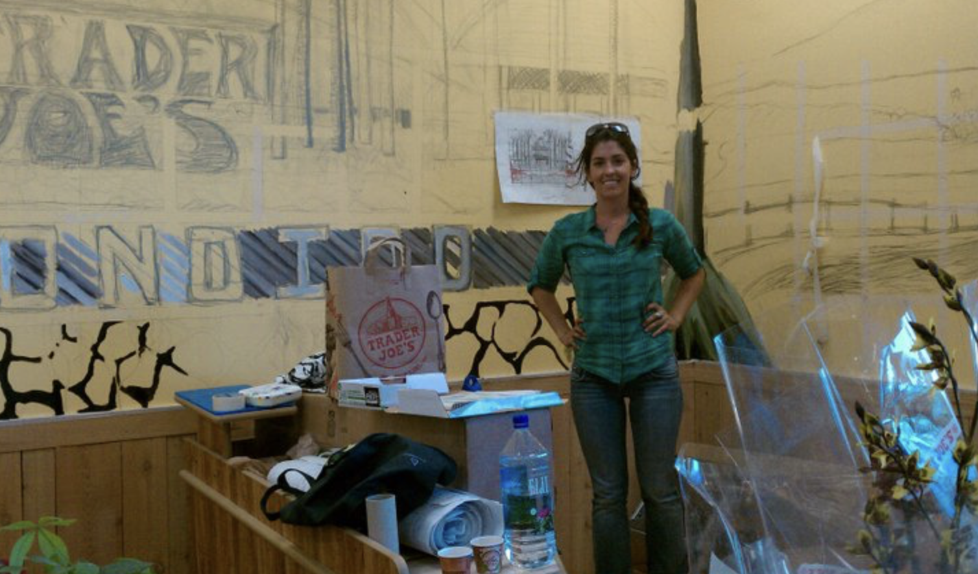 The artist standing in front of a Trader Joe's mural in progress, pencil sketch on yellow wall, 'Trader Joe's Escondido' lettering.