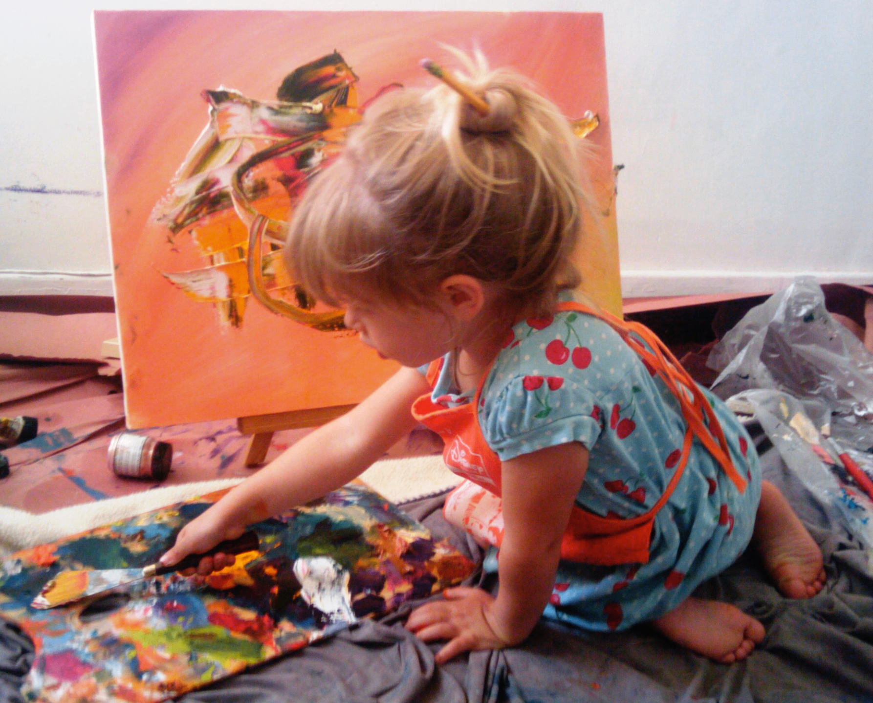 A small child mixing paints on a palette in front of her own orange abstract painting on a small easel.