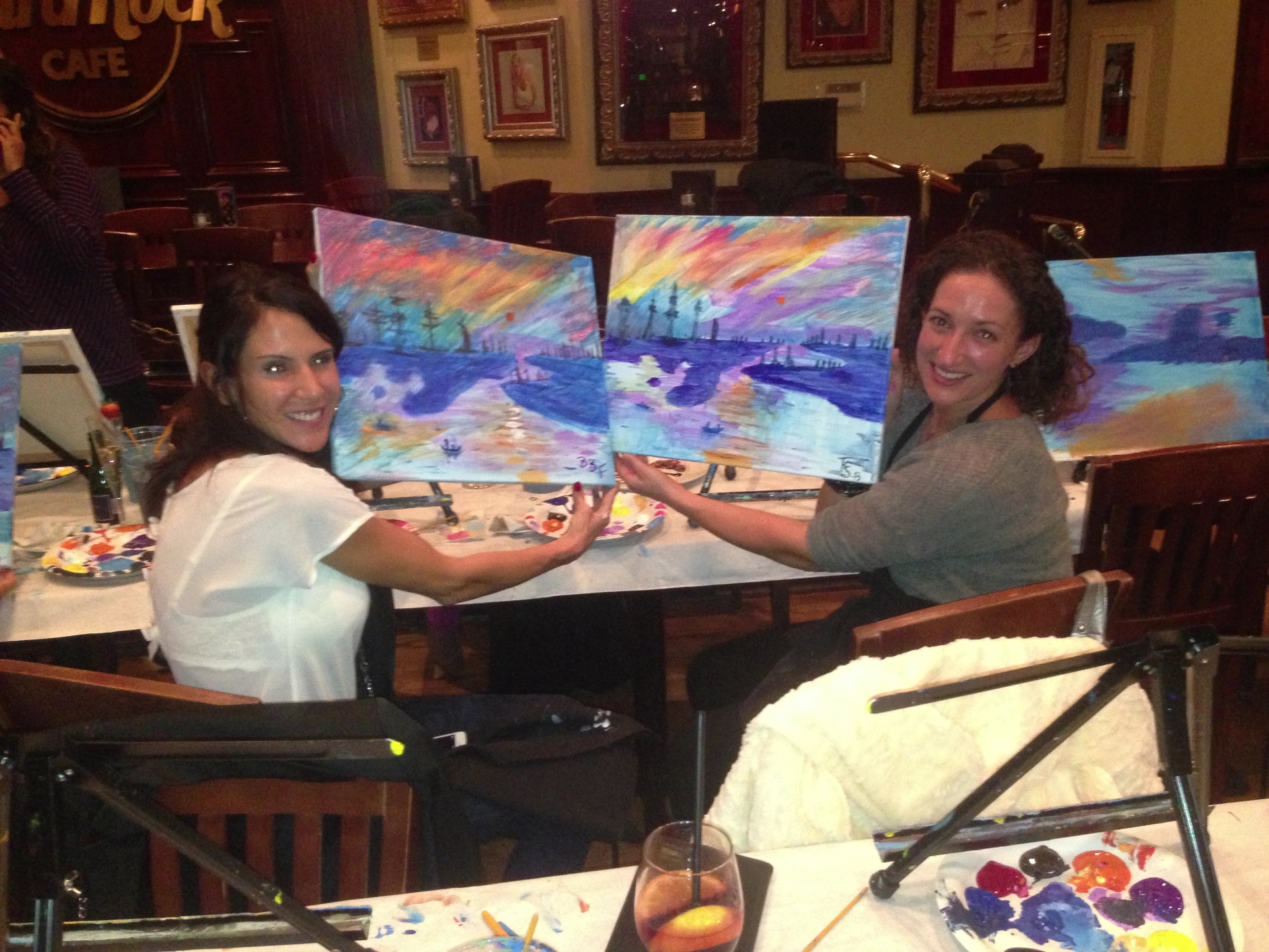 Two women holding up their finished sunset seascape paintings at a Hard Rock Cafe paint-and-sip night.
