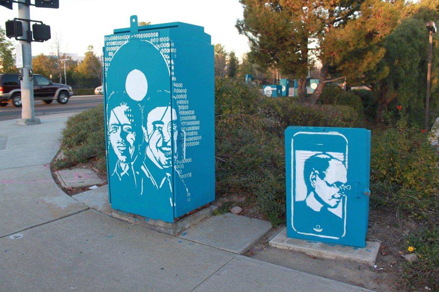 Two teal-painted electrical utility boxes on a sidewalk, one with stenciled portraits and binary, one with a smartphone silhouette framing a portrait.