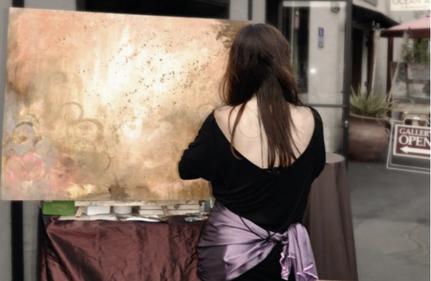 The artist painting a large gold and pink abstract canvas outside a gallery.
